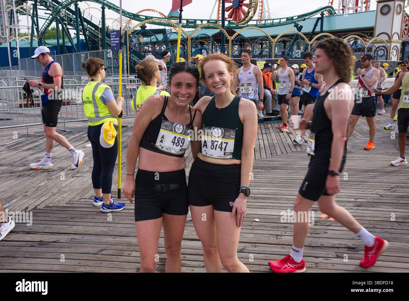 Due donne in abbigliamento da running stanno insieme su una passerella, sorridendo per una foto. Indossano bavaglino da gara. Ci sono molti altri corridori e montagne russe Foto Stock