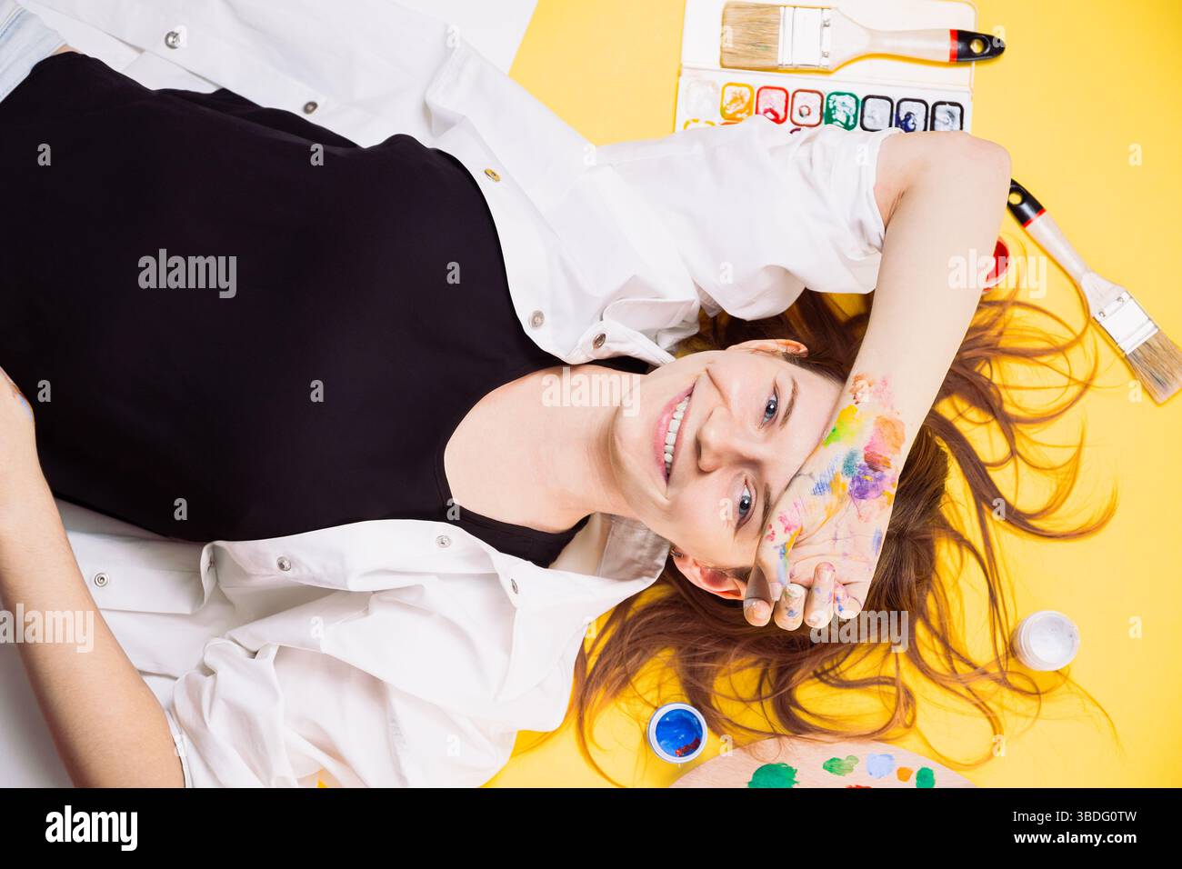 Donna allegra artista giace su sfondo giallo, dipinta a mano, circondata da pennelli e tavolozza. Studio break, stile di vita artistico, Foto Stock