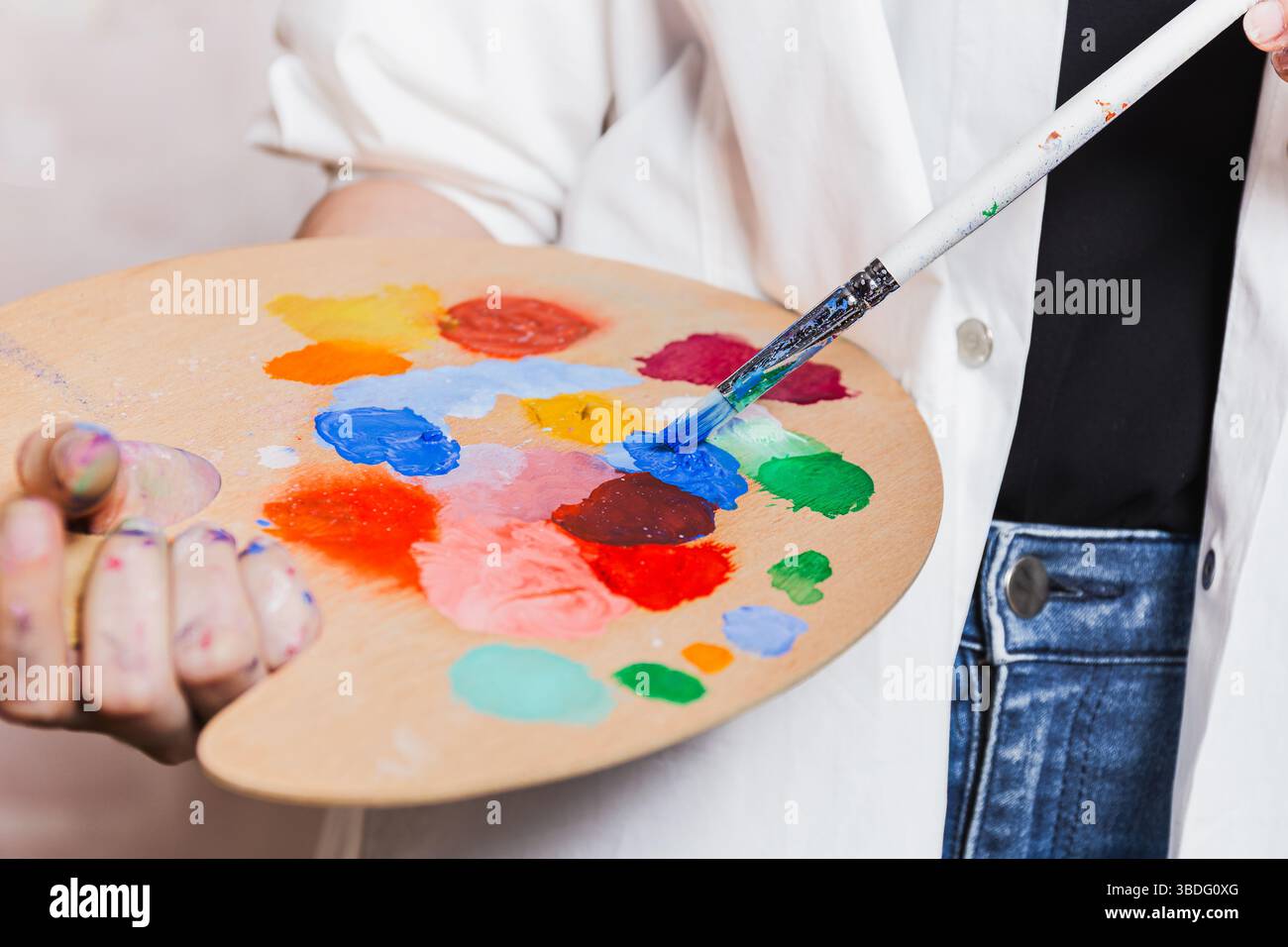 Artista che mescola la pittura su una tavolozza di legno con un pennello pieno di colori vivaci, processi creativi e strumenti artistici Foto Stock