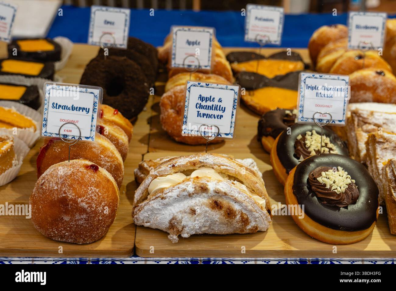 Guardando attraverso una finestra di panetteria le prelibatezze appena sfornate con una messa a fuoco selettiva. Ci sono ciambelle e fatturati con cartelli per la descrizione e i prezzi in Foto Stock