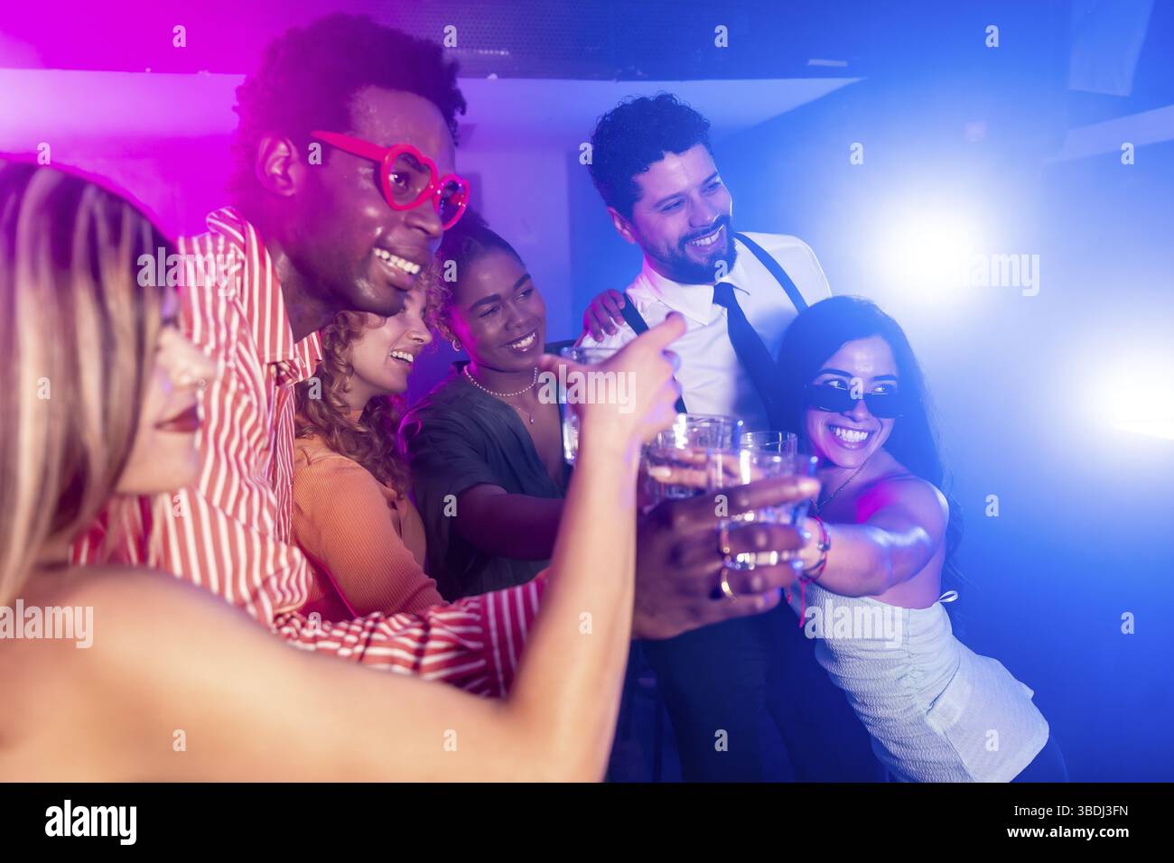 Allegri amici multietnici brindano con bevande, godendosi la vita notturna alla festa del nightclub con luci colorate Foto Stock