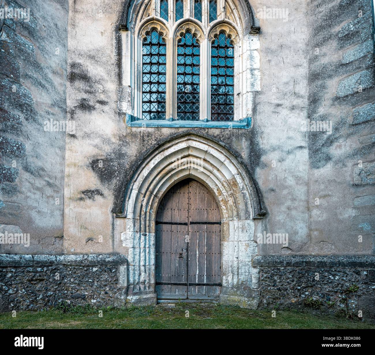 Primo piano di una pesante porta di chiesa ad arco in legno con elementi in ferro, posta sotto una finestra di vetro al piombo all'interno di un muro di pietra temprata che ha avuto Foto Stock