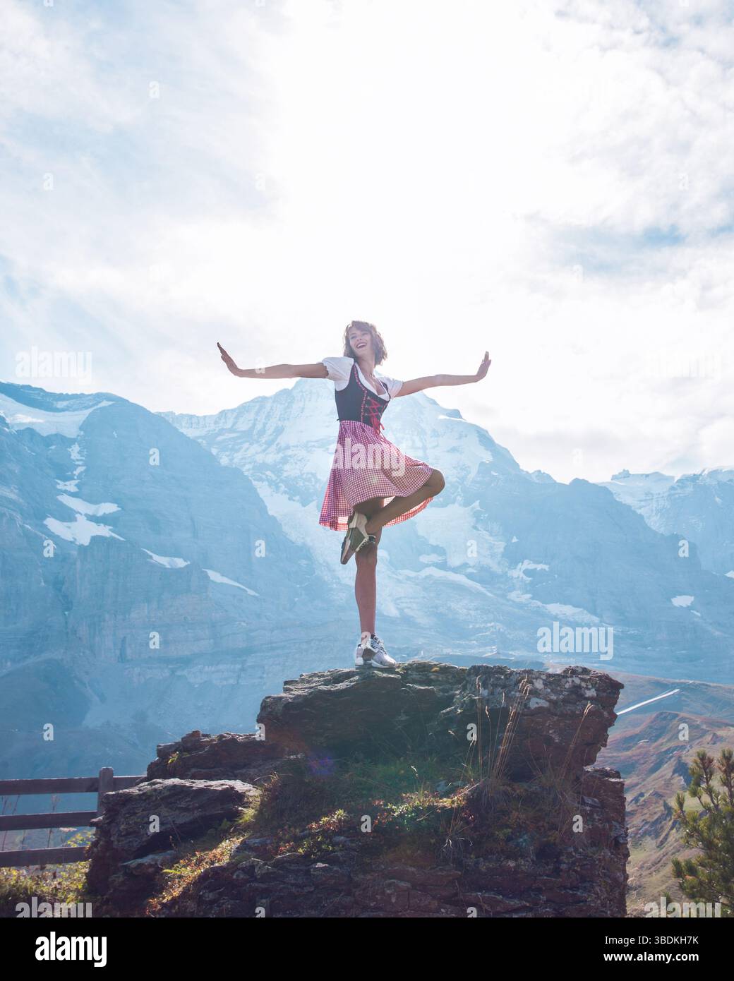Bella giovane donna che indossa abiti tradizionali bavaresi - Didndl o tracht che danzano posa carina sulla cima della roccia nel paesaggio delle Alpi europee Foto Stock
