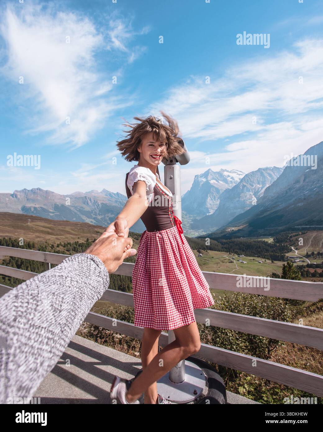 Bella giovane donna che indossa abiti tradizionali bavaresi - Didndl o tracht, tenendo la mano degli uomini dietro la macchina fotografica davanti al binoculare. Foto Stock