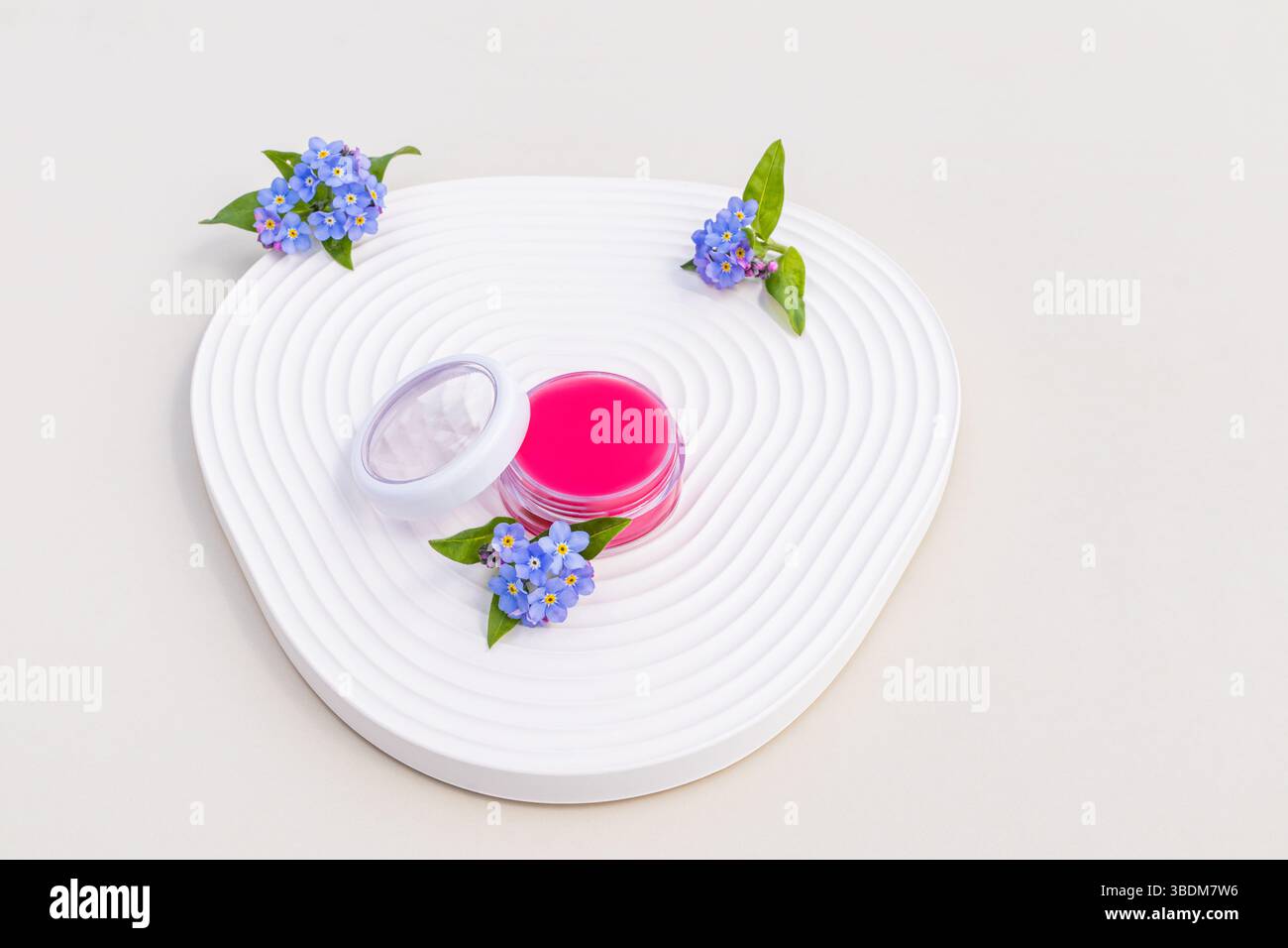 Una confezione aperta di delicato balsamo rosa si trova su un vassoio di rilievo con fiori blu. Il concetto di cura delicata, nutriente della pelle delle labbra. Foto Stock