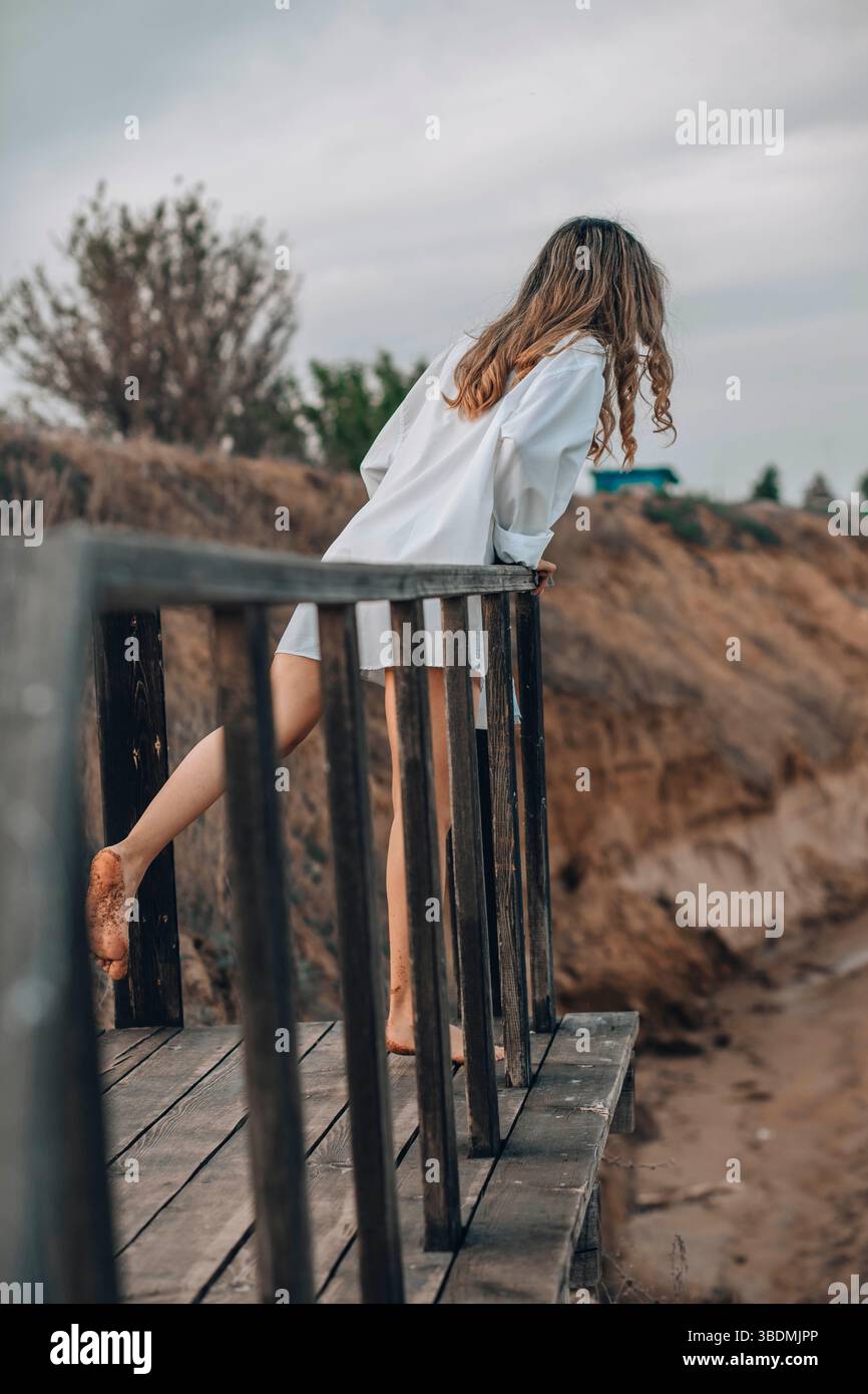 donna irriconoscibile in camicia bianca a piedi nudi su una piattaforma di legno con un piede sollevato, paesaggio naturale di scogliere, vista posteriore, solitudine, personale Foto Stock
