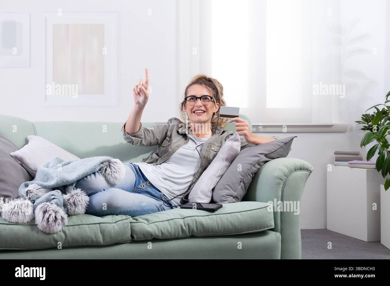 Donna sorridente sdraiata su un divano, indossa occhiali intelligenti e tiene in mano una carta di credito mentre punta verso uno schermo virtuale. Concetto di shopping online, digita Foto Stock