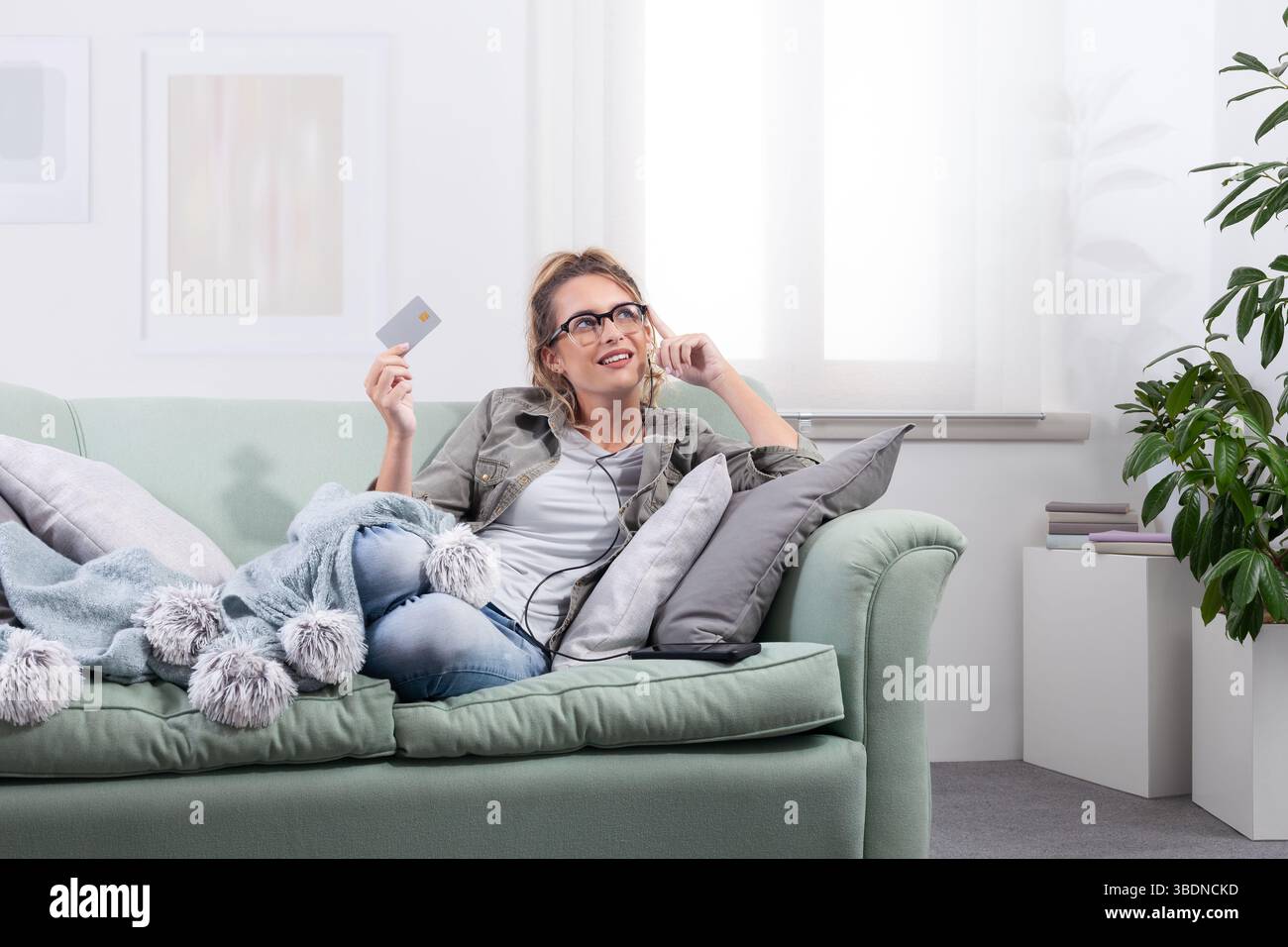 Donna sorridente sdraiata su un divano, indossa occhiali intelligenti e tiene in mano una carta di credito mentre interagisce con lo schermo virtuale. Concetto di shopping online, dig Foto Stock
