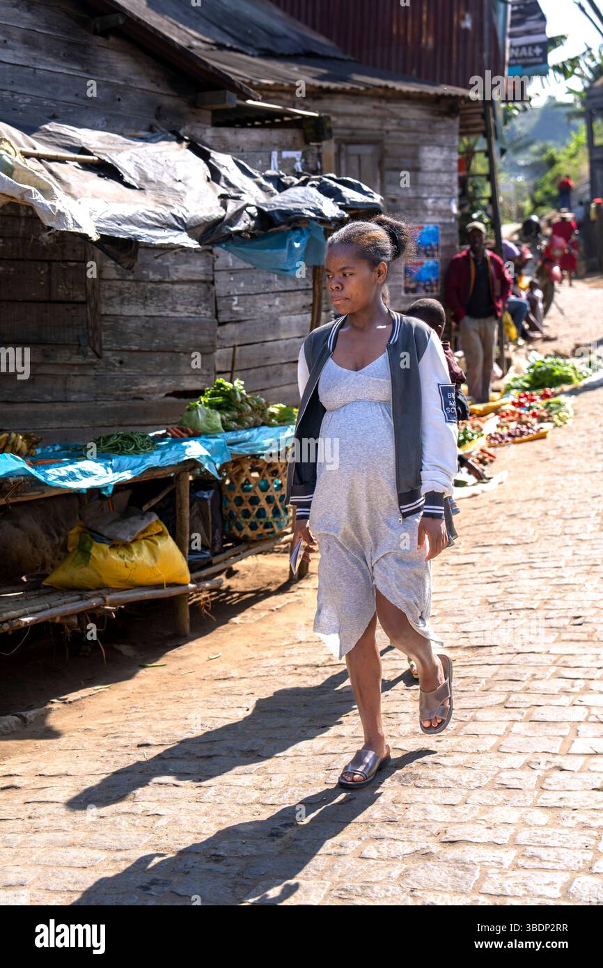 Una giovane donna malgascia con un abbigliamento casual grigio e una giacca cammina attraverso una vivace strada del mercato locale in Madagascar. Foto Stock