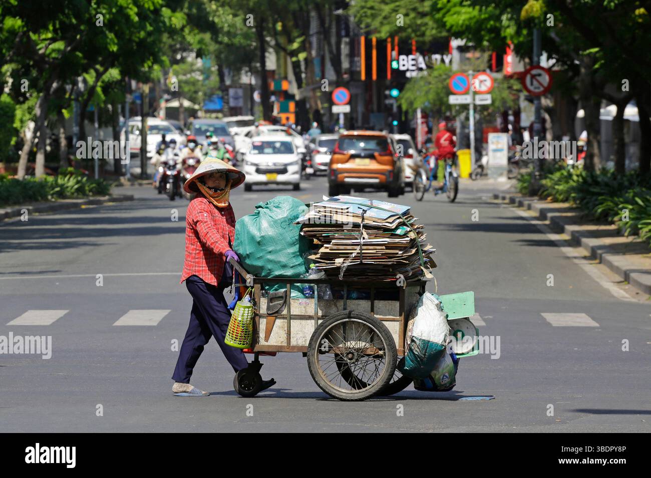 Ho chi Minh, Vietnam - 23 dicembre 2022: Donna vietnamita, che indossa un tradizionale cappello conico, non la, tira un carrello che trasporta merci per il riciclaggio. Foto Stock