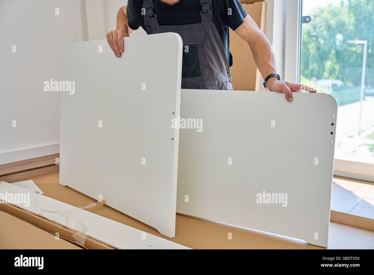 L'uomo in tuta da lavoro assembla un pannello di mobili bianco vicino a parti e utensili non imballati a casa. Tuttofare che prepara parte del letto per il montaggio in camera da letto Foto Stock