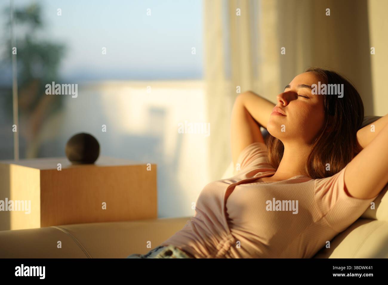 Donna rilassata che si riposa accanto a un altoparlante intelligente a casa al tramonto Foto Stock