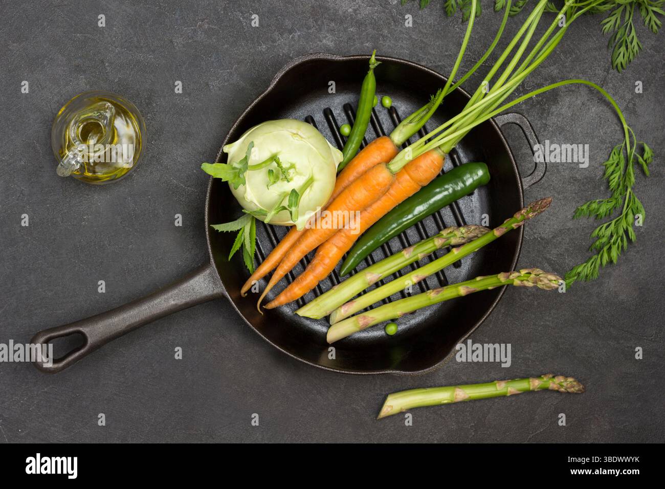 Carote fresche con cime verdi, cialda di peperone verde e asparagi in padella. Bottiglia di olio d'oliva, peperoni e piselli verdi in tavola. Sfondo nero. F Foto Stock