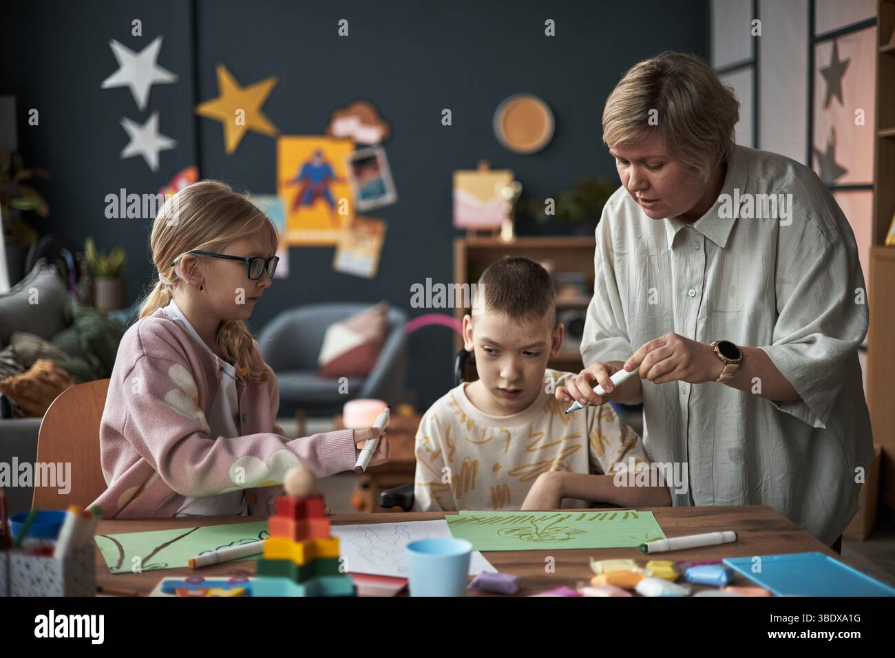 Insegnante che guida due bambini durante la lezione di arte creativa con vari materiali artigianali sul tavolo, creando opere d'arte colorate con espressioni mirate Foto Stock