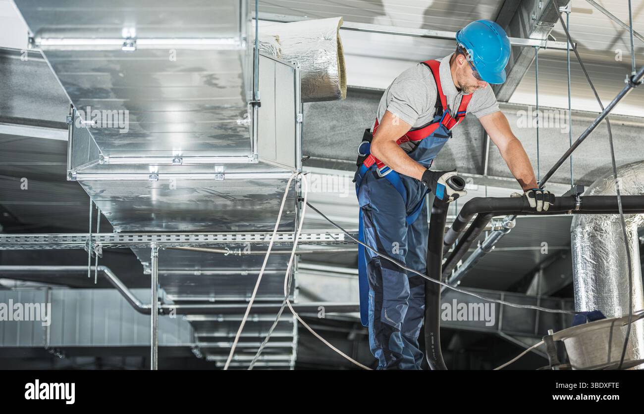 Un operatore HVAC ispeziona e ripara con attenzione un sistema di condotti dell'aria in alto in un impianto industriale, indossando dispositivi di sicurezza e concentrandosi sull'attività. Foto Stock