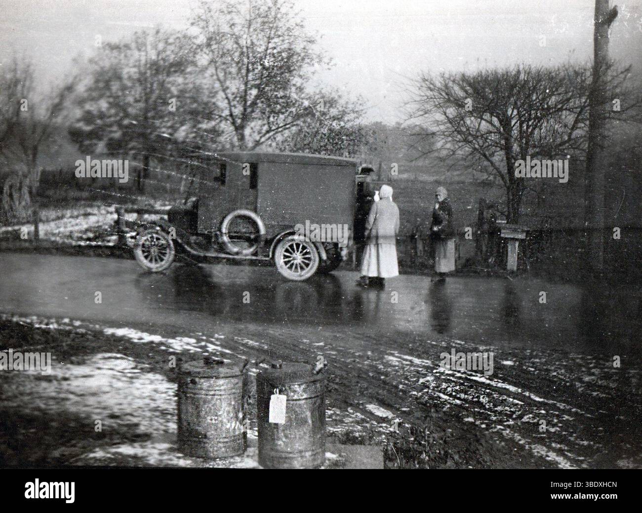 Si tratta di una foto originale e reale degli anni '1910 o '1920 che mostra una piovosa scena rurale lungo la strada. Due donne con cappotti lunghi e velo si trovano vicino a un camion per la consegna di pannelli all'inizio del XX secolo, probabilmente una Ford Model T o simili. La strada è bagnata e fangosa, con macchie di neve visibili, e una serie di lattine di latte d'epoca si trovano in primo piano, suggerendo il trasporto di latticini. Alberi senza fronzoli e una recinzione in legno aggiungono un'atmosfera rustica e ventosa. L'immagine cattura la vita quotidiana, i trasporti e le infrastrutture rurali nell'America dei primi anni del XX secolo. Foto Stock