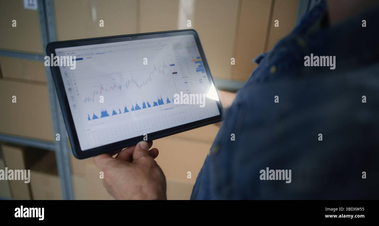 Dipendente che controlla azioni in tempo reale e grafici Exchange Market utilizzando un computer tablet digitale. Uomo che effettua investimenti durante l'orario di lavoro in magazzino, combinando lavoro e Trading online. Primo piano. Foto Stock