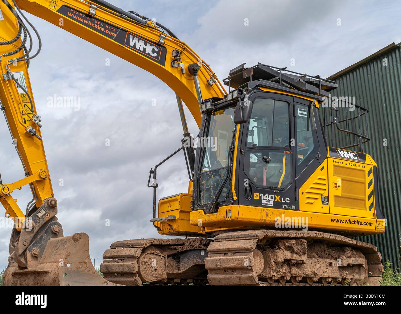 Vista laterale ravvicinata di una scavatrice meccanica JCB a riposo di fronte a un edificio di una fattoria in acciaio, nel Regno Unito, con cieli nuvolosi sopra. Foto Stock