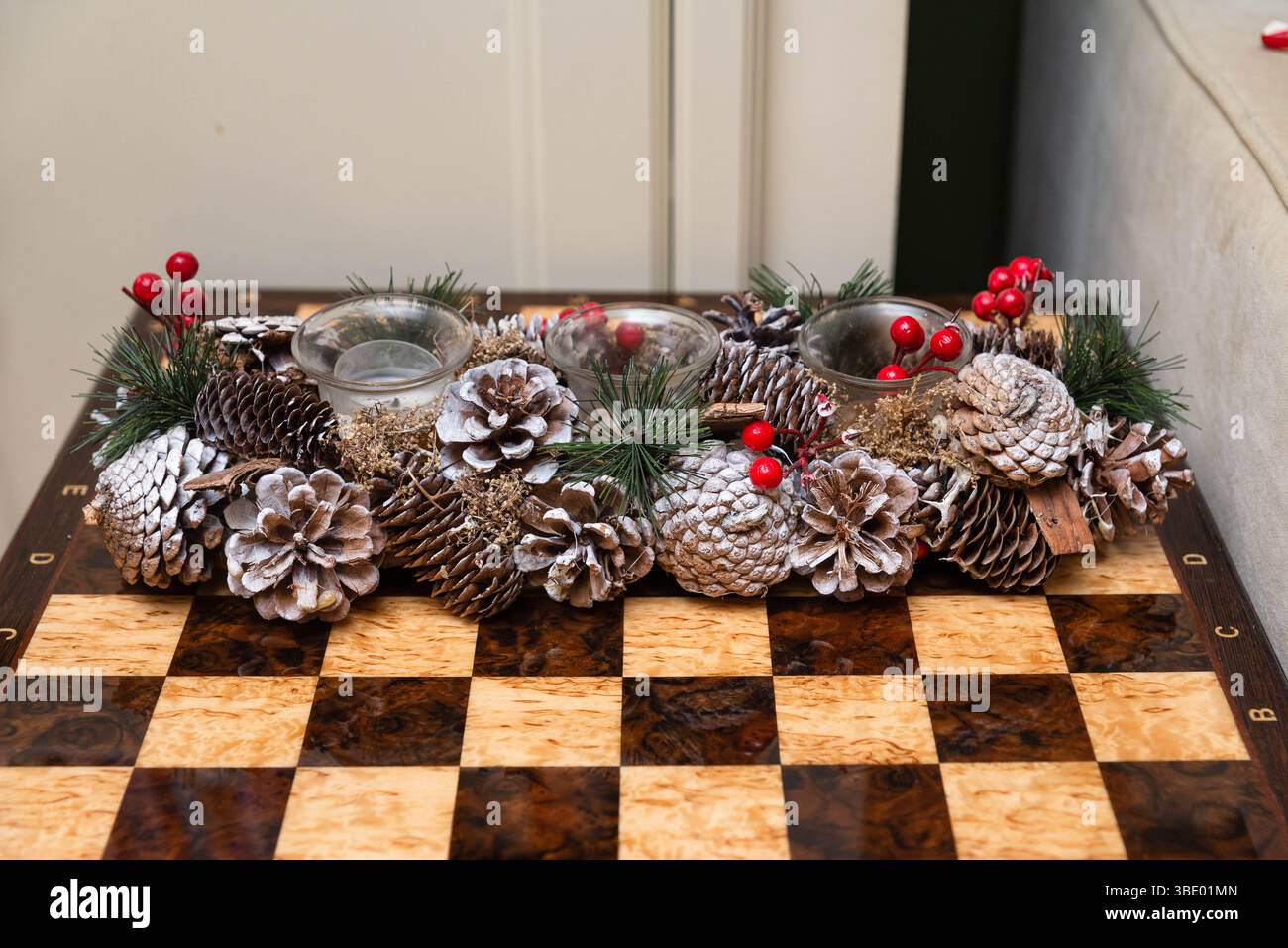 Elemento centrale del portacandele a tema natalizio con pinne, frutti di bosco e vegetazione smerigliati esposti su un tavolo decorativo a scacchiera in legno. Foto Stock