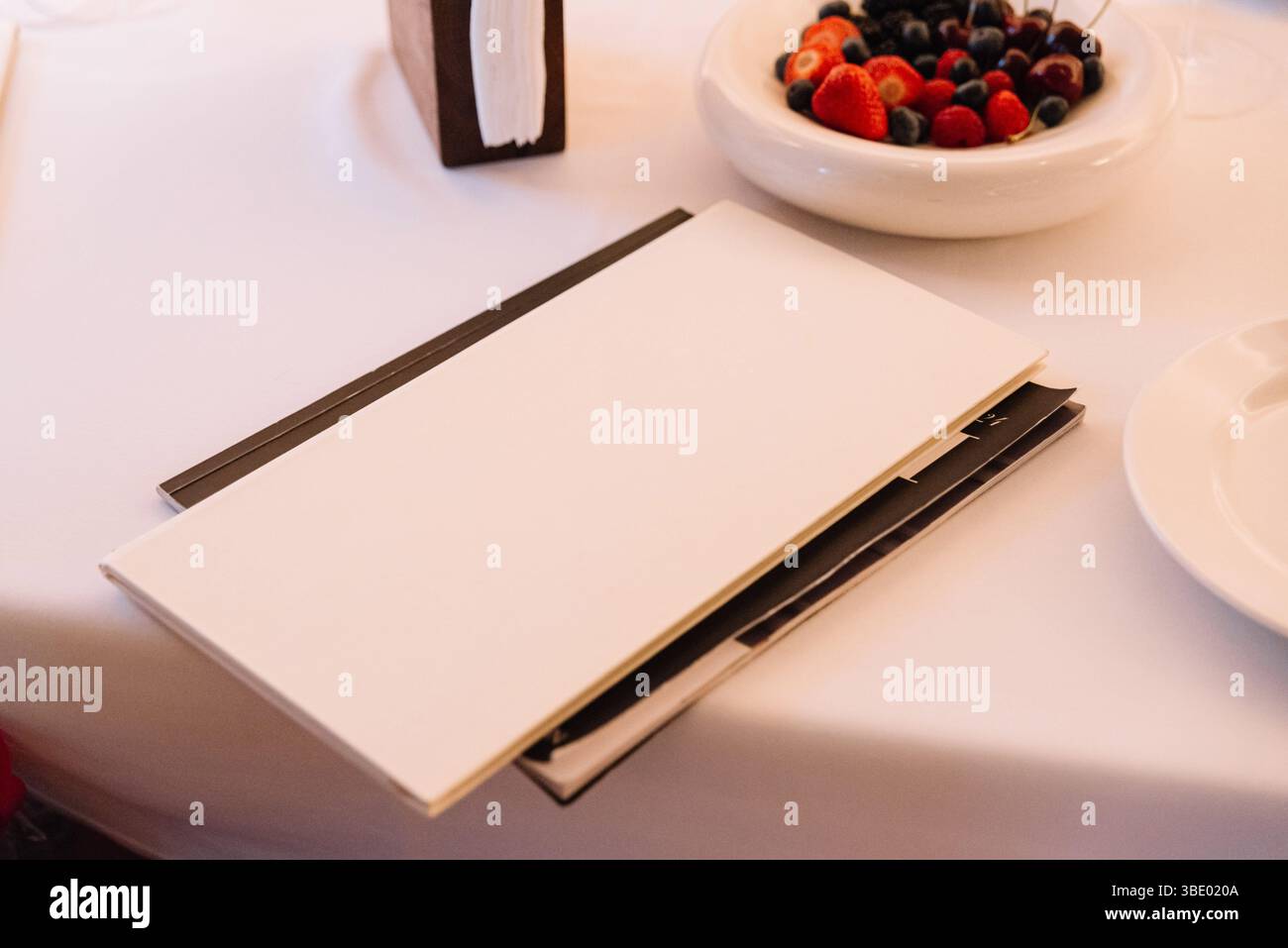 Elegante ambiente da tavola in un ristorante con menu bianco, portasciugamani e una ciotola di frutti di bosco e ciliegie fresche assortite. Foto Stock