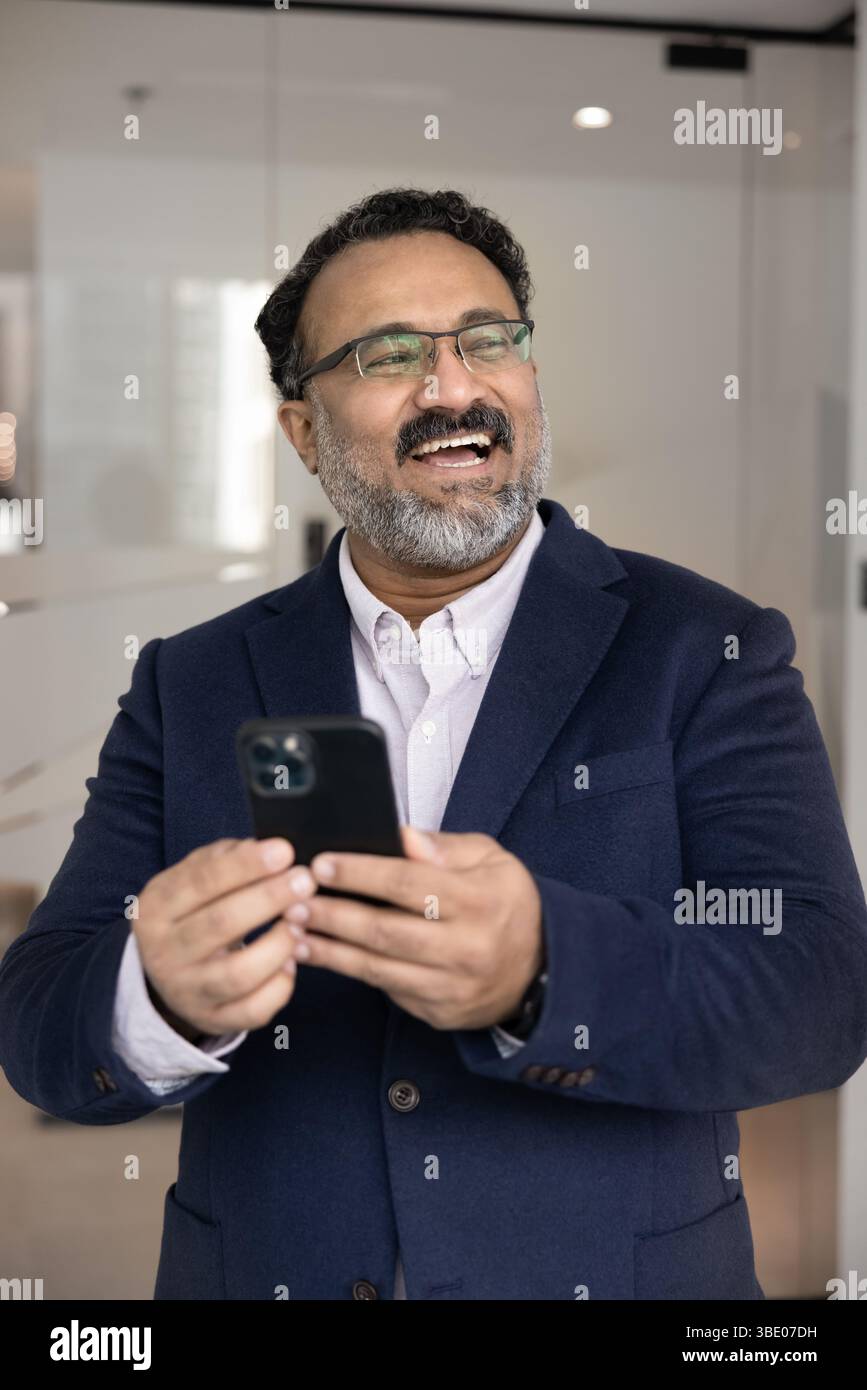 Felice uomo d'affari indiano in tuta in piedi sul posto di lavoro con smartphone Foto Stock