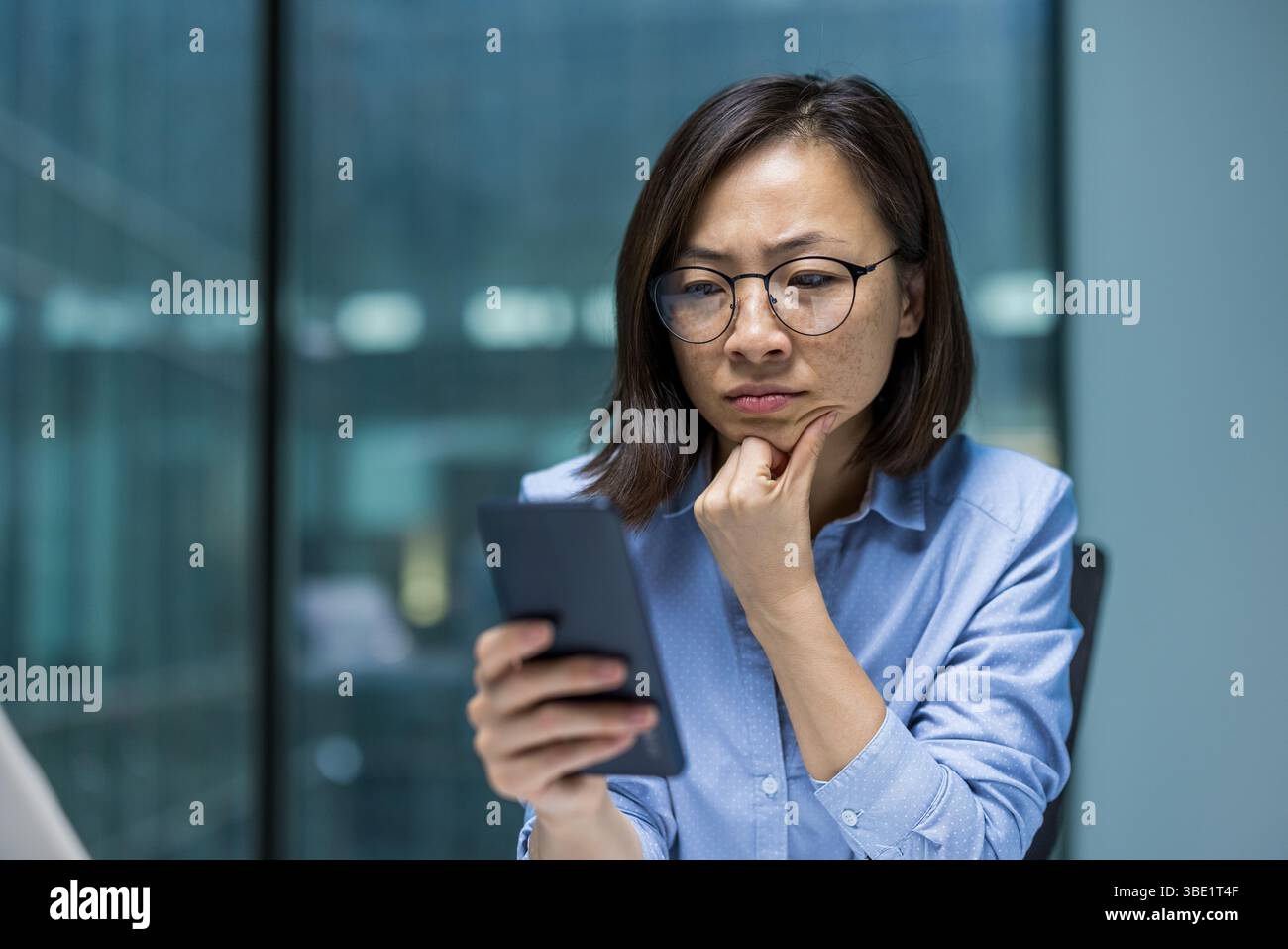 Una donna asiatica premurosa che indossa gli occhiali esamina il suo smartphone, riflettendo su un messaggio in un moderno ambiente d'ufficio. Foto Stock
