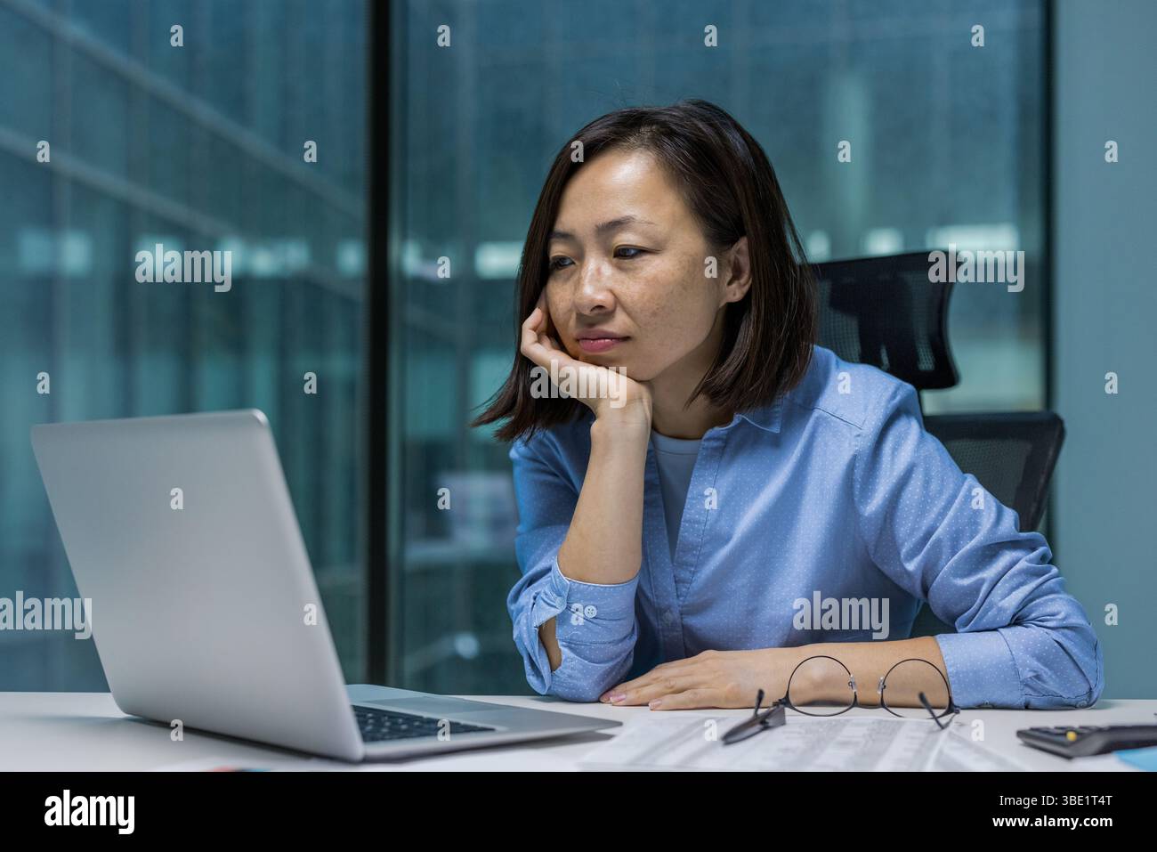 Una donna asiatica penosa siede alla sua scrivania, guardando lo schermo del suo notebook con un'espressione premurosa, forse di notte. Foto Stock