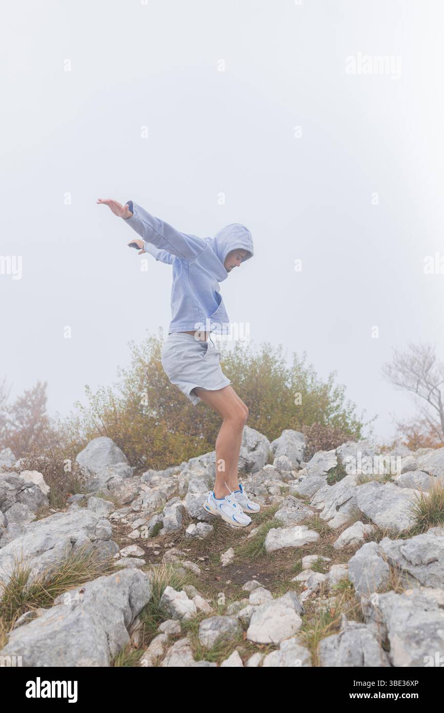 L'uomo tocca il terreno dopo il salto con la nebbiosa pendenza rocciosa indossando felpa con cappuccio e pantaloncini sportivi. Foto Stock