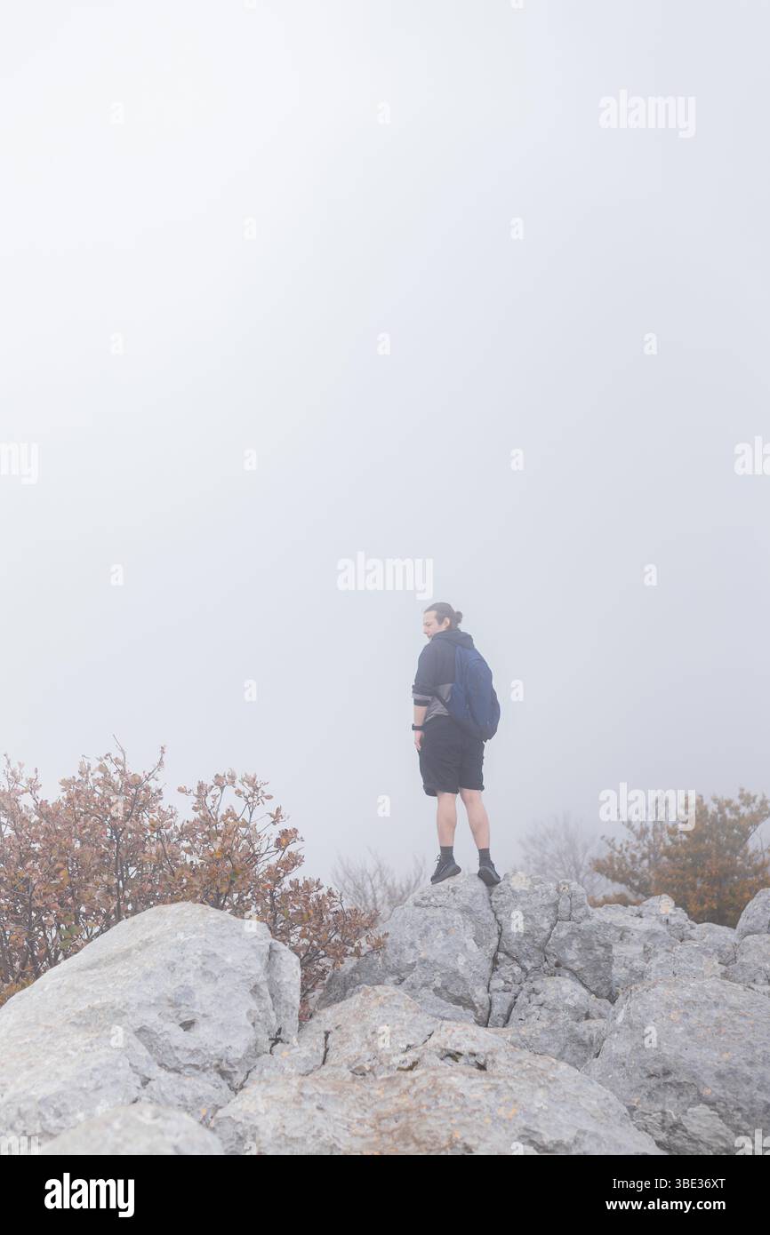 L'uomo si erge in cima alla cresta rocciosa delle montagne indossando un abbigliamento sportivo circondato da fitta nebbia e arbusti autunnali. Foto Stock