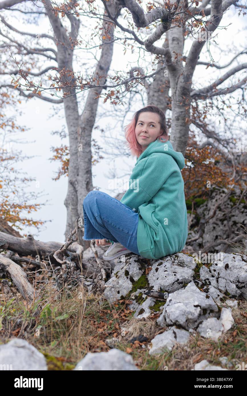 Donna con capelli rosa e felpa con cappuccio verde seduta tranquilla su un terreno roccioso sotto gli alberi autunnali. Foto Stock