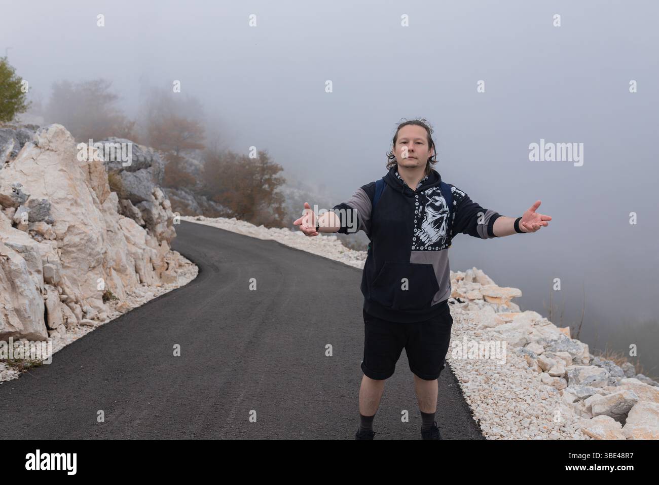 L'uomo apre le braccia in piedi su una strada di montagna nebbiosa circondata da scogliere Foto Stock