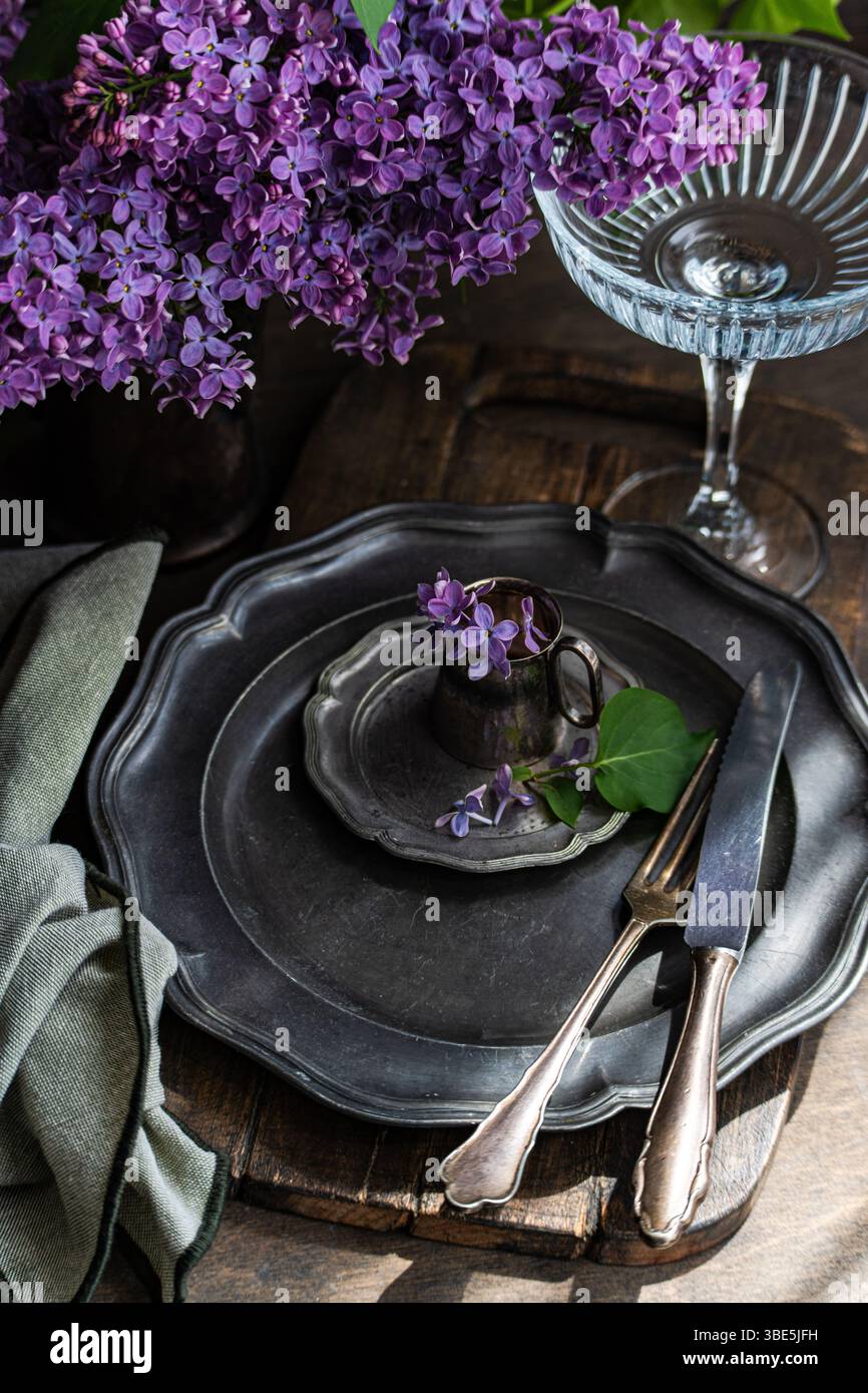 Un ambiente rustico da tavolo presenta piatti e posate di peltro antichi, adornati con vibranti fiori lilla in una tazza vintage. Un tovagliolo pieghettato e un bicchiere aggiungono Foto Stock