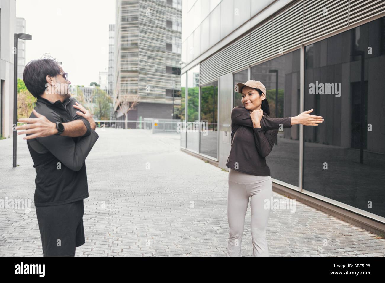 Coppia che indossa un abbigliamento sportivo che si estende all'aperto in un ambiente urbano. Si preparano per una giornata attiva con una routine di riscaldamento, che mette in mostra fitness e guarigione Foto Stock