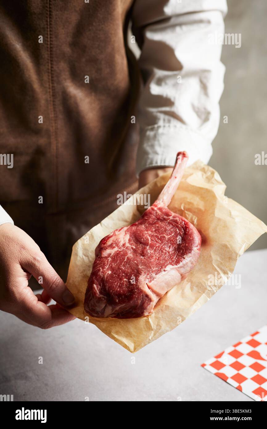 Primo piano di un macellaio o di uno chef che tiene una bistecca di tomahawk cruda avvolta in carta da macelleria marrone. Il taglio spesso e osseo è ideale per grigliare o per un'esperienza gastronomica Foto Stock