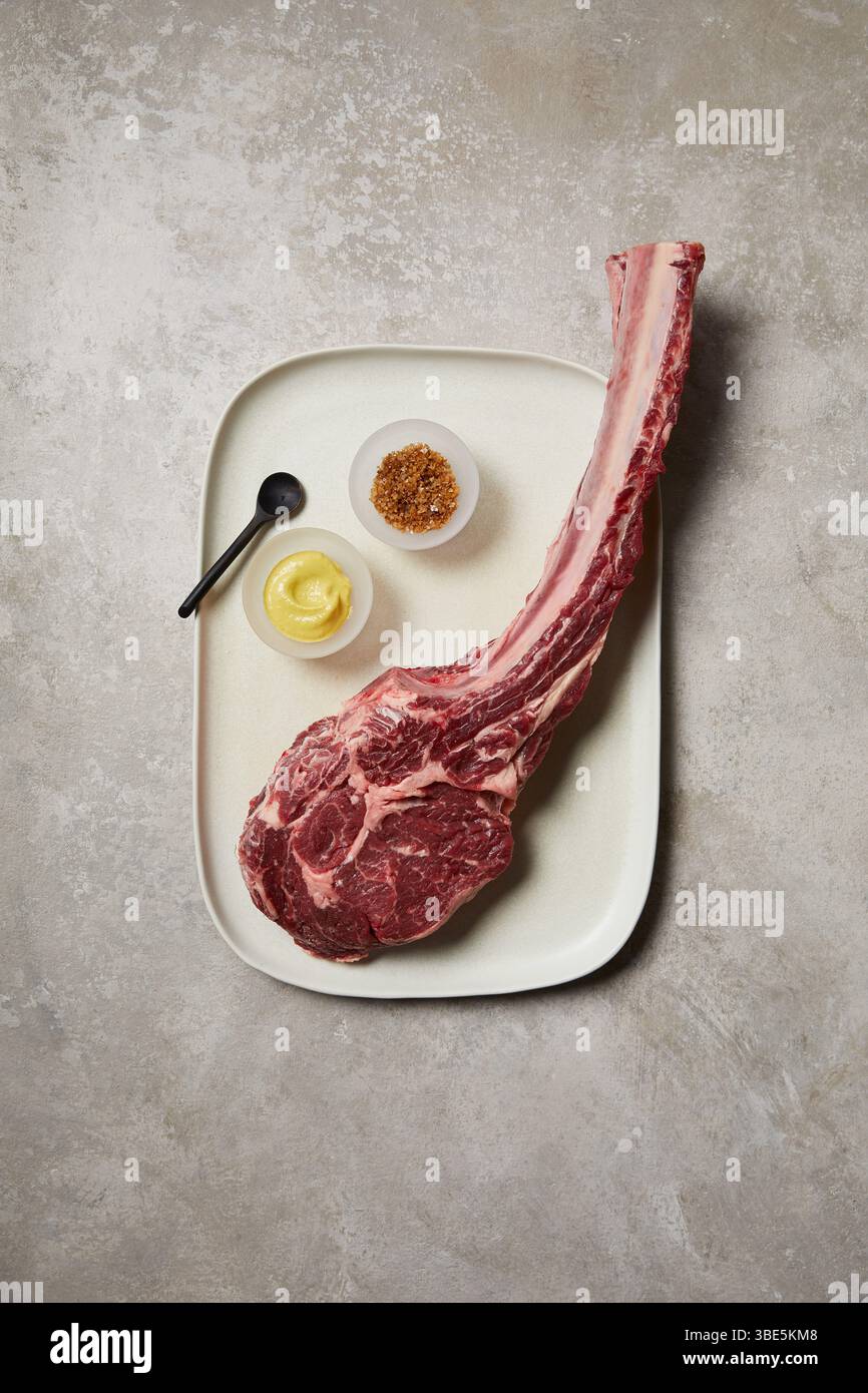 Vista dall'alto verso il basso di una bistecca di tomahawk cruda presentata su un vassoio di ceramica, accompagnata da una piccola ciotola di senape, spezie e un cucchiaio nero. Un taglio di qualità superiore Foto Stock