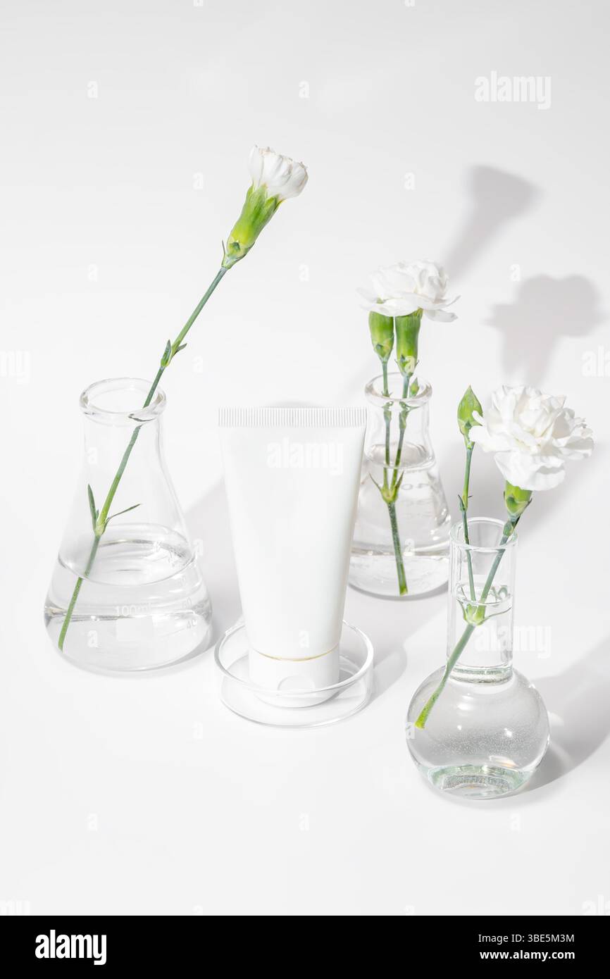 Tubo bianco per la cura della pelle tra fiasche da laboratorio con fiori in fiore, simbolo di prodotti di bellezza organici e ricerca scientifica in cosmetici. Foto Stock