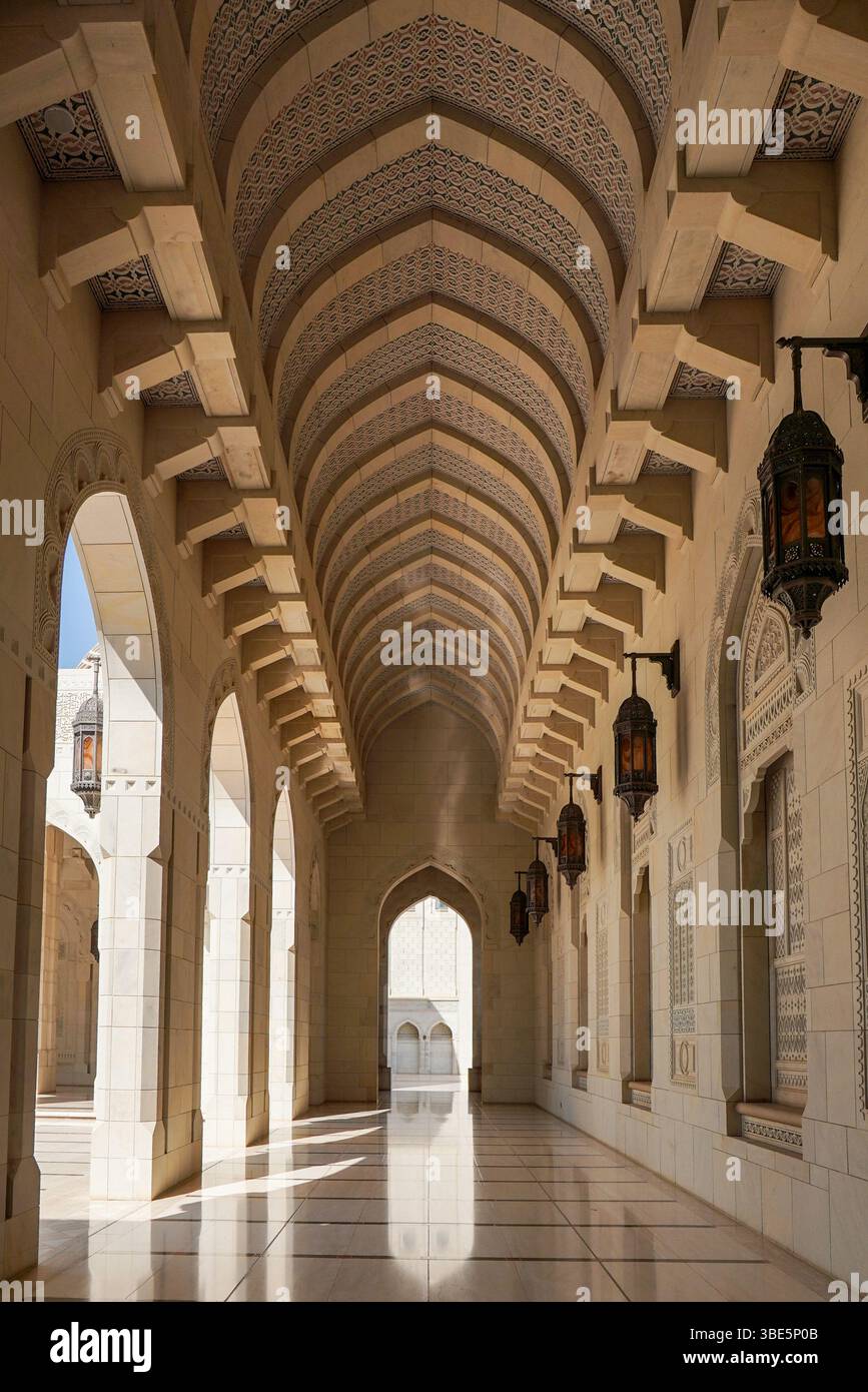 Eleganti arcate e intricati motivi adornano il corridoio della Moschea del Sultano Qaboos a Mascate, Oman. La luce del sole scorre attraverso, gettando ombre sui poli Foto Stock