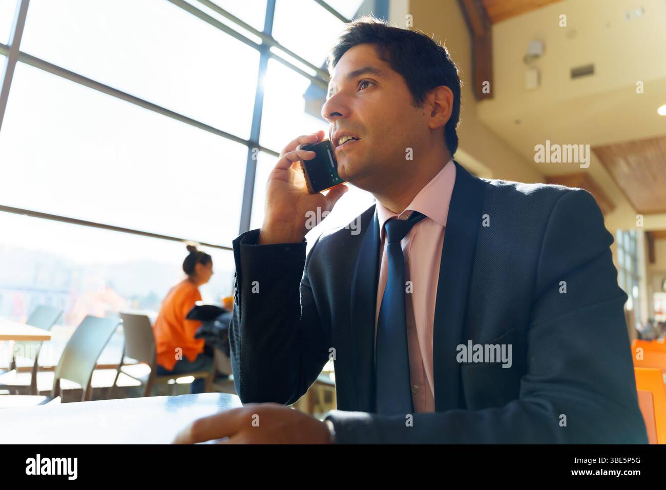 Un giovane uomo d'affari cileno, vestito con una tuta, parla al telefono in un ambiente moderno e soleggiato con grandi finestre, concentrandosi sulla conversazione Foto Stock