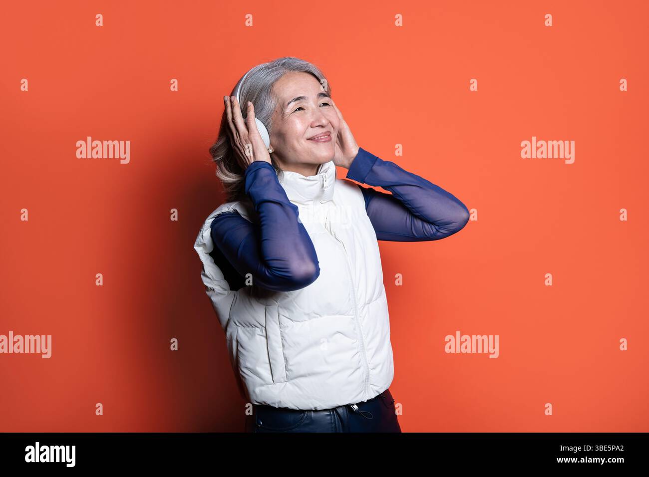 Donna asiatica cinese anziana che indossa le cuffie, sorride e ascolta musica. Vestito con un gilet bianco e maniche blu, appoggiato su un dorso arancione Foto Stock