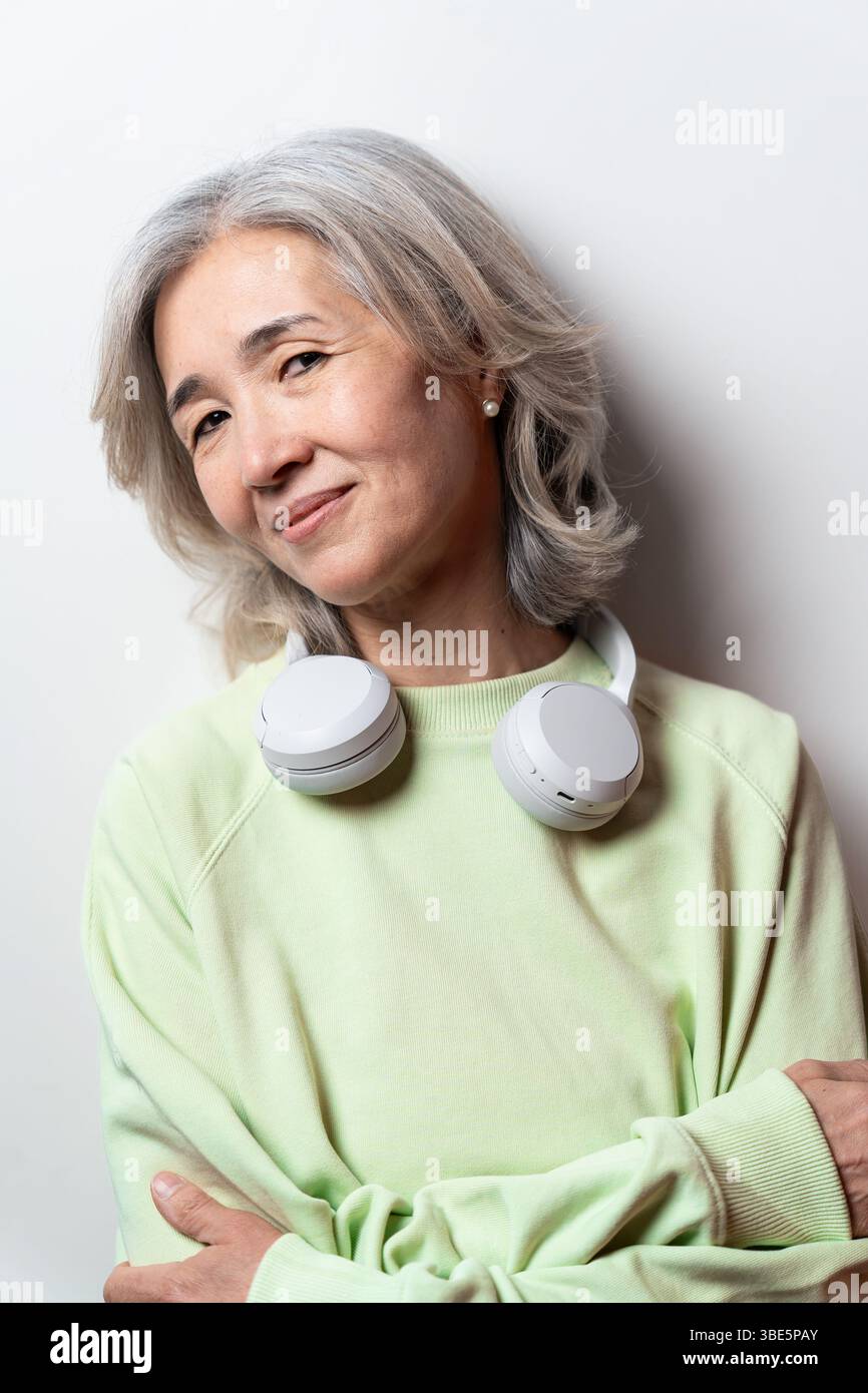 Donna asiatica cinese anziana con capelli grigi, indossa un maglione verde chiaro e cuffie bianche intorno al collo, che guarda con fiducia la fotocamera Foto Stock