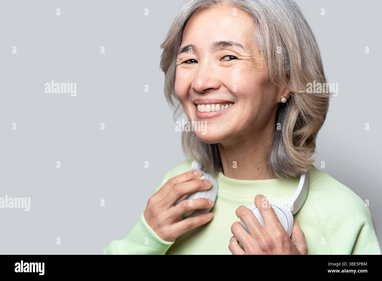 Donna asiatica cinese anziana con i capelli grigi che sorride calorosamente, indossa un maglione verde chiaro, tiene le cuffie bianche e goditi la sua esperienza musicale i Foto Stock