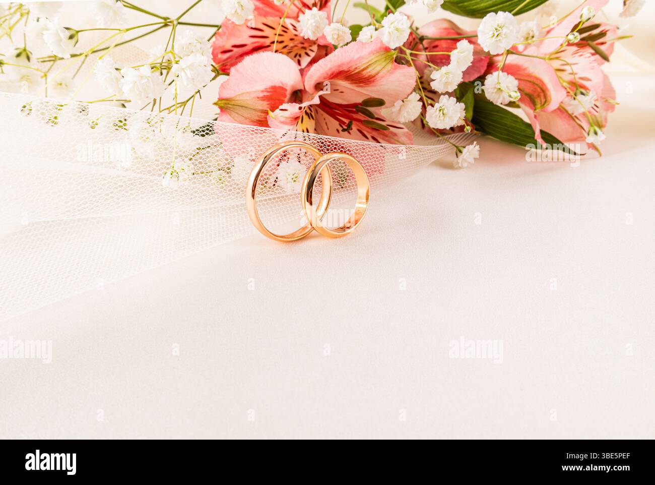 Elegante composizione di due fedi nuziali in oro circondate da delicati fiori rosa e bianchi su una superficie bianca soffice, a simboleggiare l'amore e l'impegno Foto Stock