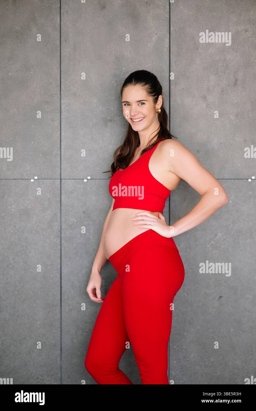 Donna incinta che sorride con un vestito da yoga rosso, in piedi con sicurezza contro una moderna parete grigia. Abbracciare il benessere della maternità e il fitness in un ambiente confortevole Foto Stock