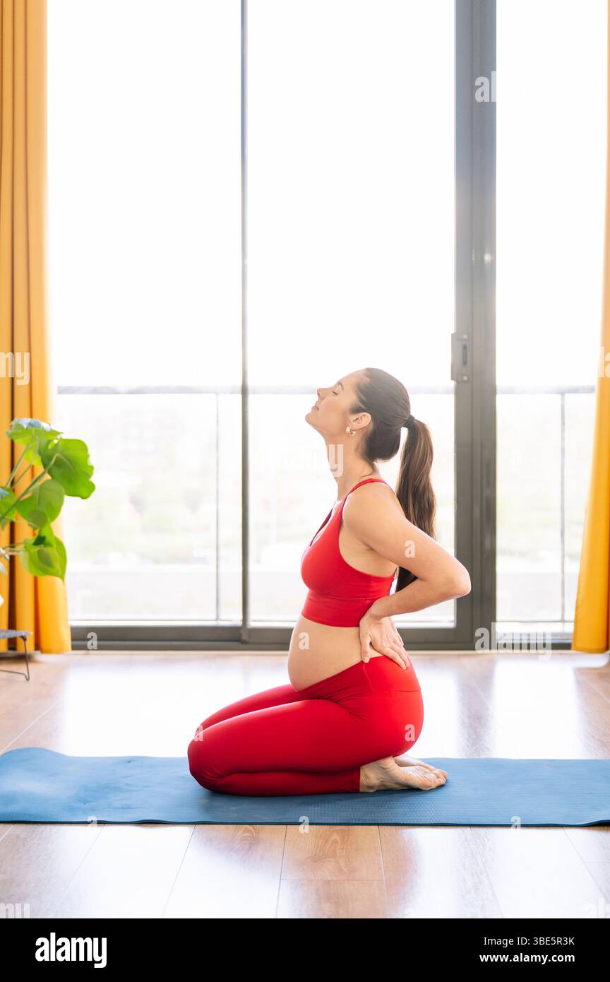 Una donna incinta pratica yoga in un ambiente sereno, abbracciando benessere e relax. Si inginocchia su un tappetino da yoga in un abbigliamento sportivo confortevole, promuovendo Foto Stock