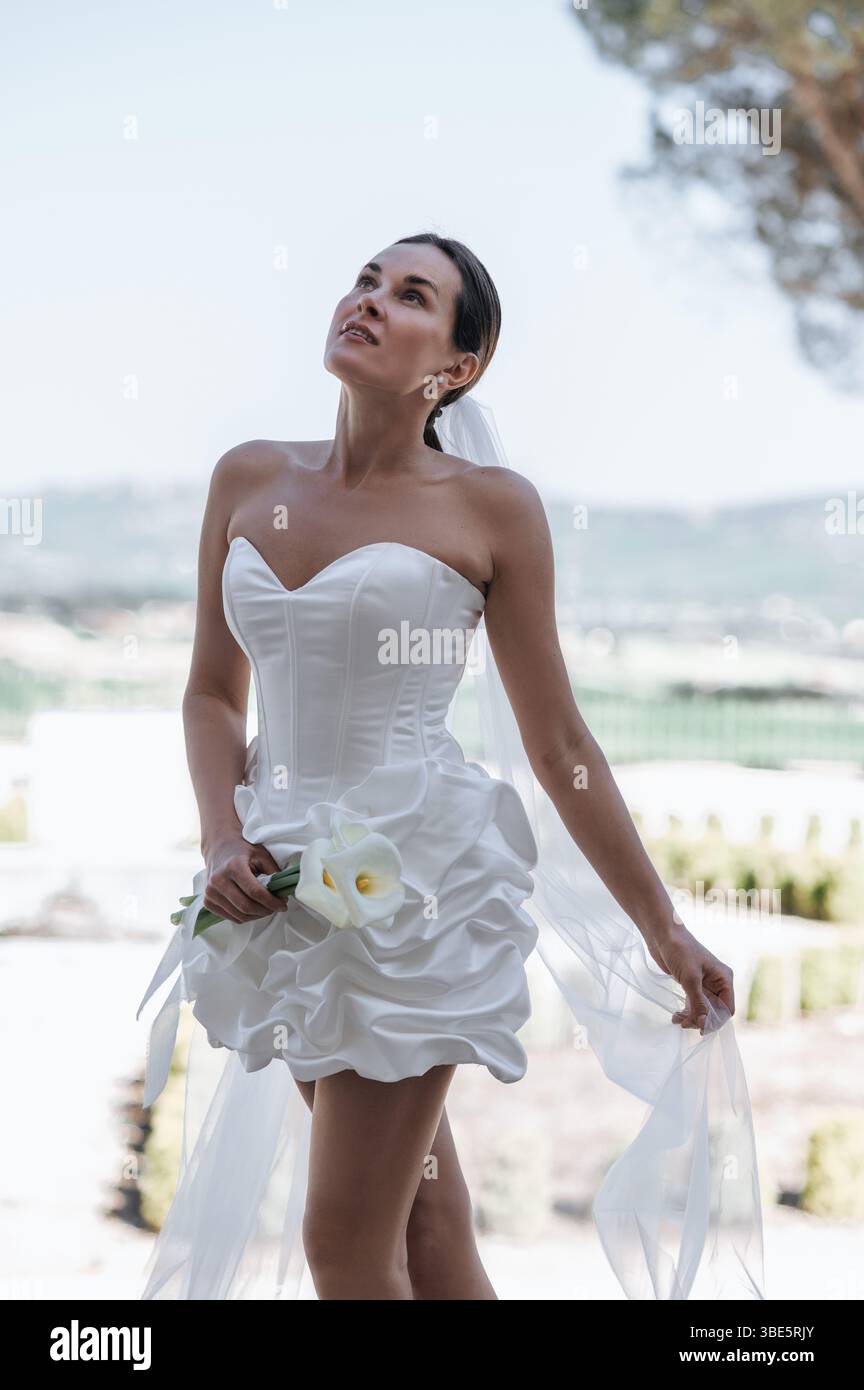 Una splendida sposa in un abito senza spalline sorge elegantemente nel giardino di un'antica villa a Treia, Macerata. Lo sfondo panoramico esalta la bellezza di Foto Stock