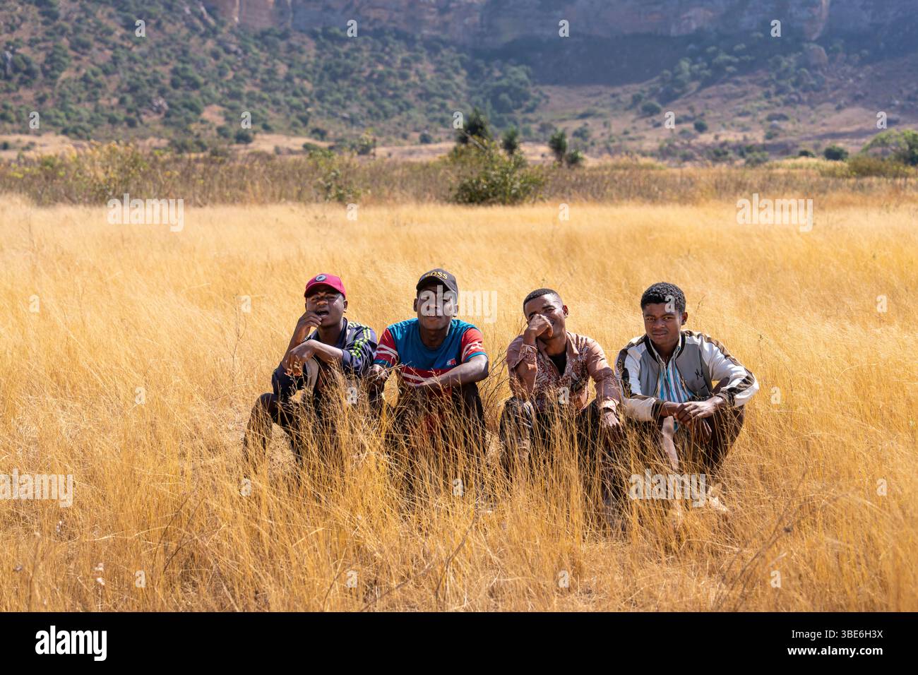 Quattro giovani malgasci siedono in un campo di erba dorata con aspre colline sullo sfondo, godendosi il sole e il paesaggio aperto. Foto Stock