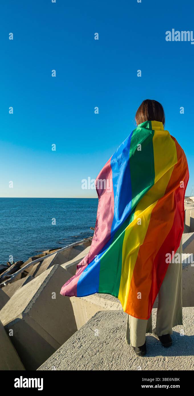 Giovane donna sulla schiena su un molo di fronte al mare, avvolta da una bandiera LGTBI. La bandiera copre gran parte del corpo del modello. La luce naturale fa risaltare il Foto Stock