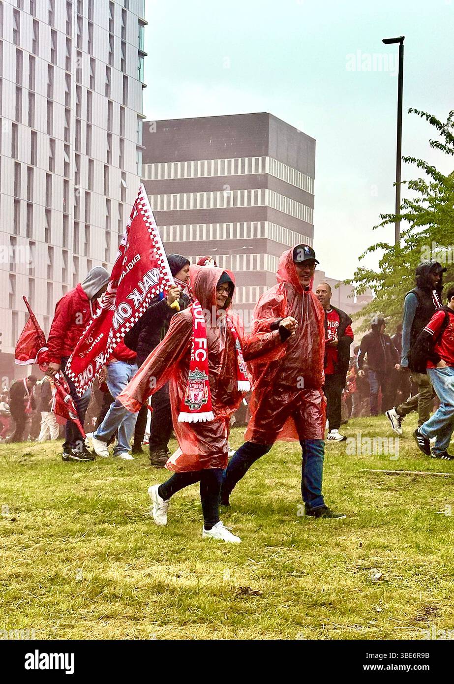 Una coppia che si tiene per mano in poncho rossi con una grande bandiera su Hunter Street, Liverpool, Regno Unito, 26 maggio 2025, durante le celebrazioni per la vittoria della Premier League. - Immagine stock catturata con smartphone