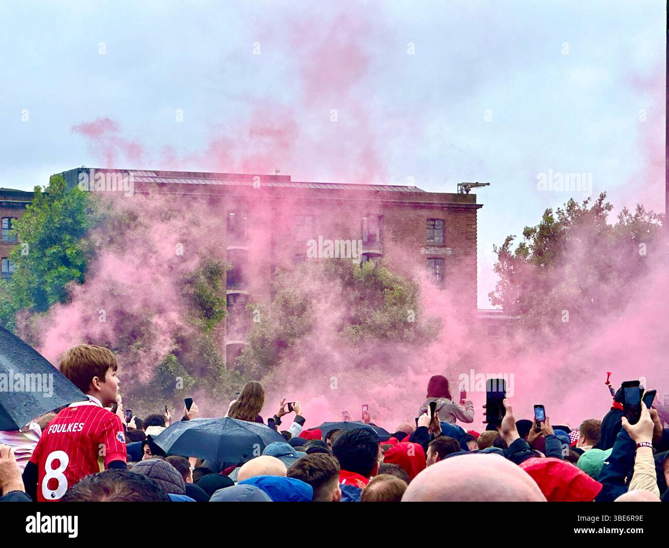 I tifosi con ombrelli e telefoni sollevati attendono la parata della vittoria del Liverpool FC a Strand Street, Liverpool, Regno Unito, 26 maggio 2025. Il fumo rosso riempie l'aria. - Immagine stock catturata con smartphone