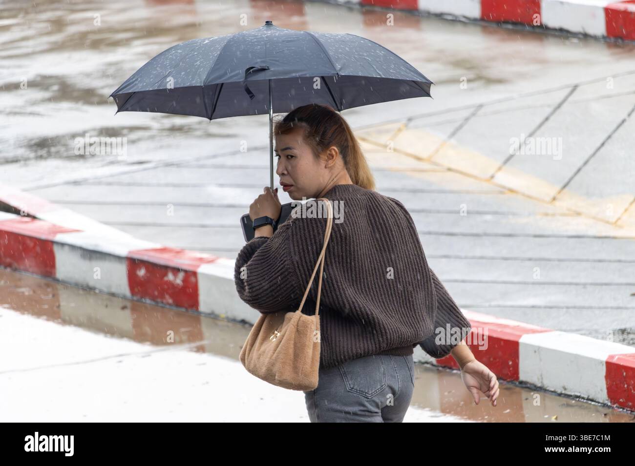 SAMUT PRAKAN, THAILANDIA, 16 MAGGIO 2025, Una donna che porta un ombrello in una giornata di pioggia passeggia accanto a una strada con marciapiedi dipinti di rosso e bianco Foto Stock